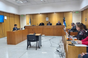 Una sala de audiencias con varias personas en mesas de madera, monitores, banderas, micrófonos y cables en el suelo