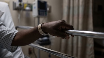 Mano de persona con piel oscura sosteniendo baranda de cama de hospital. Se ve un brazo con bata, pulsera de identificación y equipo médico desenfocado de fondo.