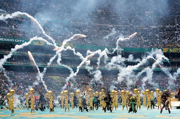 En un Stadium, artistas con trajes dorados y penachos actúan. Fuergos artificiales y humo blanco se elevan sobre la multitud de aficionados