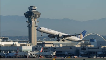 Un avión blanco de United Airlines con motores gemelos despega con el tren de aterrizaje extendido. Detrás, la torre de control de LAX y montañas