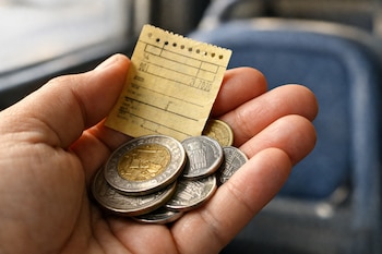 Mano sosteniendo un boleto de bus amarillo pálido y un puñado de monedas metálicas. Un asiento de colectivo azul oscuro aparece desenfocado al fondo.