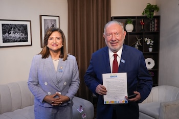 Un hombre con traje azul y una mujer con traje azul claro posan de pie. Ambos llevan un pin de bus azul. El hombre sostiene un documento con texto