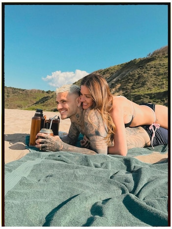 Pareja sonriente tumbada en una toalla verde sobre la arena de la playa. El hombre sostiene un mate. Fondo de colinas verdes y cielo azul