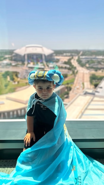 Un niño posa frente a un ventanal, viste una camiseta negra, una capa celeste y un sombrero con el escudo de AFA. Detrás hay un estadio y una carretera