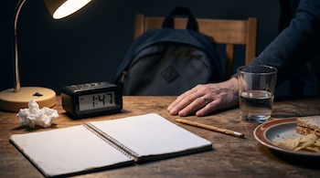 Escritorio de madera con una lámpara encendida, un reloj digital, un cuaderno abierto, un lápiz, un vaso con agua, un plato de comida y una mano.