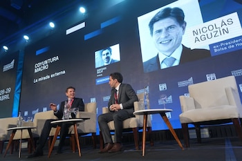 Dos hombres en trajes sentados en sillas de tela en un escenario, con mesas y botellas de agua. Pantallas grandes muestran nombres de ponentes y el título del evento