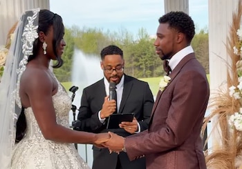 Una pareja se toma de las manos frente a un oficiante durante su boda al aire libre. La mujer usa un vestido blanco y velo, el hombre un traje marrón. Detrás hay una fuente