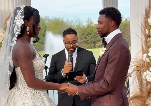 Noah Lyles y Junelle Bromfield se toman de las manos durante su ceremonia de boda al aire libre, oficiada por un ministro, con una fuente de agua de fondo. (Captura de un video publicado por @junellebromfield en Instagram)