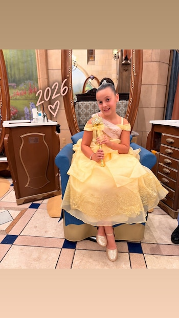 Niña vestida con un vestido amarillo de princesa, una tiara y un cetro dorado, sentada en un sillón azul frente a un espejo