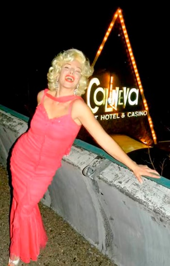 Una persona con peluca rubia y vestido rosa brillante posa de lado sonriendo frente a un letrero de neón que dice "Cal Neva HOTEL & CASINO" en la noche