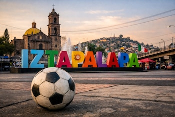 Bola de futebol em primeiro plano em terreno rochoso, em frente às letras coloridas de Iztapalapa, igreja colonial e colina com população ao pôr do sol.