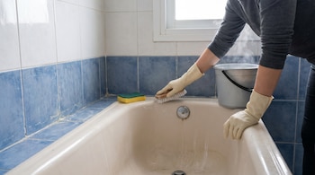 Una persona con guantes beige frota el interior de una bañera color crema con un cepillo, junto a un balde con agua caliente humeante y una esponja.