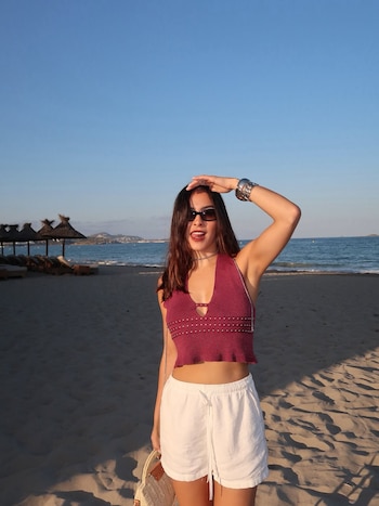 Una mujer con pelo largo oscuro, gafas de sol, crop top color vino, shorts blancos y sandalias está de pie en una playa arenosa junto al mar