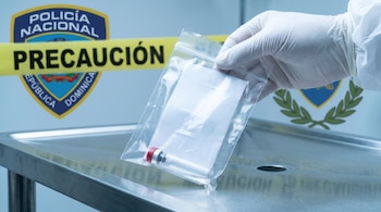 Mano enguantada sostiene una bolsa de evidencia transparente con un frasco y papel. Fondo difuso muestra cinta 'Precaución' y escudo Policía Nacional Dominicana.