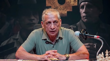Hombre de mediana edad con cabello gris y polo verde, sentado ante un micrófono, con manos entrelazadas. De fondo, gráficos y silueta de rostro