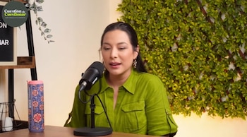 Natalia Salas, una mujer de cabello oscuro y blusa verde, habla en un micrófono en un estudio de podcast con una pared cubierta de follaje verde y un termo decorado
