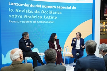 Tres ponentes, dos hombres y una mujer, sentados en un escenario azul y amarillo. Un hombre habla por micrófono. La pantalla muestra el título del evento. Público en primer plano