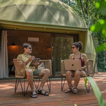 Dos personas, un hombre y una mujer, sentadas en sillas de mimbre en una terraza de madera, usando una tableta y una computadora portátil frente a un domo verde