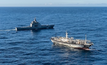 Vista aérea de un buque de guerra gris con el número 53 en su casco, navegando a la izquierda de una embarcación blanca con plataforma en el mar azul