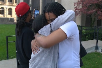 Un hombre con trenzas abraza a una persona de espaldas mientras otro individuo graba con un teléfono frente a un edificio