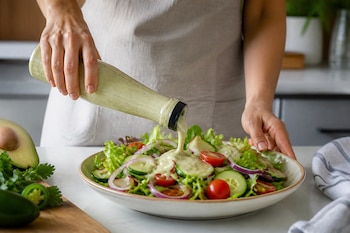 Manos de una persona vertiendo aderezo verde desde una botella sobre un plato de ensalada con lechuga, tomate, cebolla y pepino.