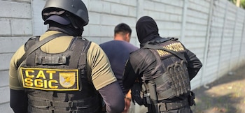 Tres agentes de policía escoltan a un hombre esposado que viste una camisa roja y pantalones cortos grises, junto a un vehículo policial y árboles
