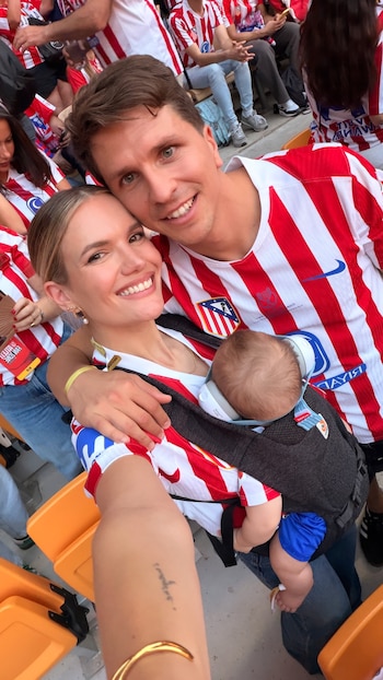 Gianluca Simeone y Eva Barigela sonriendo, con camisetas del Atlético de Madrid, y su bebé Faustino en un portabebés con auriculares en un estadio