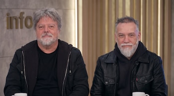 Dos hombres con cabello y barba canosos, vestidos con chaquetas oscuras, sentados. Un logo que dice "info" en la pared de fondo. Hay dos tazas blancas.