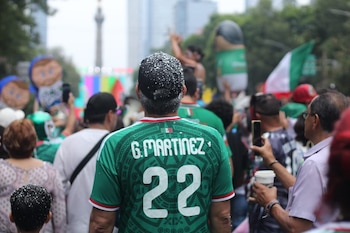 Multitud de personas de espaldas. Hombre en primer plano con camiseta verde de fútbol, número 22 y gorra. Ángel de la Independencia y bandera mexicana al fondo