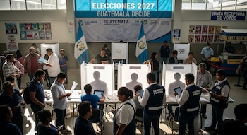 Interior de un centro con cabinas de votación y personas, incluyendo funcionarios, bajo una pancarta azul que anuncia "ELECCIONES 2027 GUATEMALA DECIDE".
