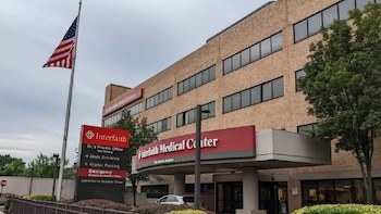 Vista exterior del Interfaith Medical Center, un edificio de ladrillo de varios pisos con ventanas oscuras, un letrero rojo, una bandera de EE. UU. y árboles
