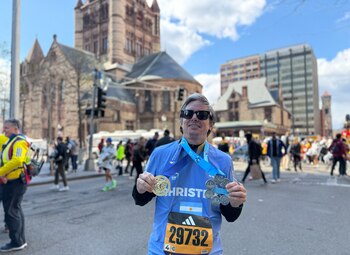 Christian Asinelli, un hombre de mediana edad con gafas de sol, sonríe y muestra medallas de maratón con una camiseta azul del número 29732; se ve gente y edificios al fondo