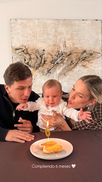 Eva Bargiela y Gianluca Simeone con su bebé Faustino. Una vela "6" encendida y pastelitos en la mesa, frente a un cuadro abstracto
