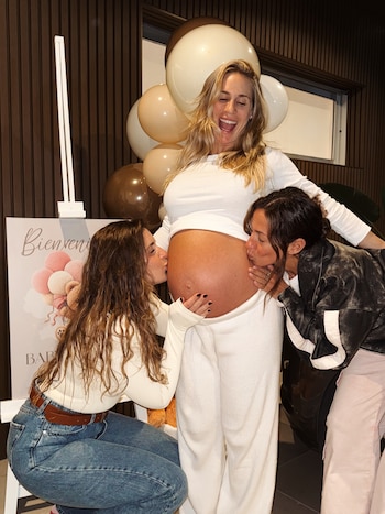 Una mujer embarazada rubia sonríe, vestida de blanco, mientras dos mujeres besan su vientre. Globos beige y un cartel de "Bienvenida" se ven de fondo