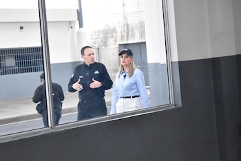 Grupo de personas con ropa formal y uniformes militares en el pasillo de una prisión flanqueado por celdas con rejas y personas en el fondo