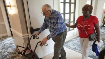 Un anciano con camisa a cuadros azul y andador rojo avanza, asistido por una mujer con camiseta roja. Se observa un reloj Citizen en su muñeca izquierda