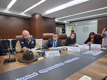 Una reunión de varias personas sentadas alrededor de una mesa de madera. Hay micrófonos, documentos y placas de identificación con nombres y organizaciones. Un sombrero está sobre la mesa