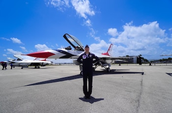 Piloto sonriente con uniforme oscuro posa frente a un jet de combate blanco, rojo y azul en pista de asfalto. Jets adicionales al fondo bajo cielo azul con nubes