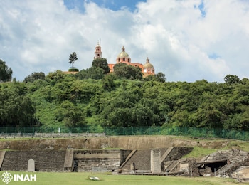 Estructuras de piedra de una pirámide, un cerro cubierto de vegetación y un templo colonial con cúpulas y torre en la cima