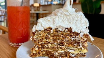 Primer plano de una alta porción de torta de capas con relleno de dulce de leche y cobertura de merengue blanco, en plato blanco, con bebida roja al fondo