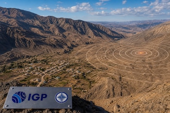 Paisaje montañoso con un poblado rural, ondas concéntricas sobre el terreno y una placa metálica en primer plano con el logotipo de IGP.