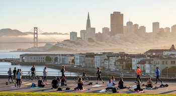 Vista panorámica de San Francisco con el puente Golden Gate y rascacielos al fondo; en primer plano, varias personas practican yoga y corren al aire libre.