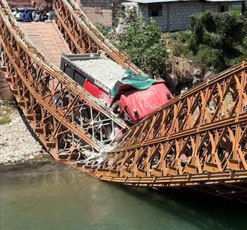 Un puente metálico colapsado sostiene un camión volquete de color rojo cargado con piedras sobre un río