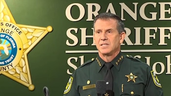 Hombre de mediana edad con uniforme verde de sheriff y corbata negra, con insignias doradas, hablando frente a un micrófono. De fondo, el logo y el texto de 'ORANGE SHERIFF'