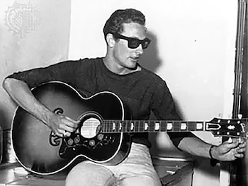 Buddy Holly viste gafas de sol oscuras y camiseta negra mientras sostiene una guitarra acústica y ajusta una clavija