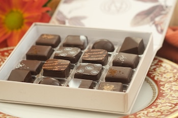 Una caja abierta contiene dieciséis bombones de chocolate de diferentes formas y decoraciones. Un plato decorativo rojo y dorado se observa al fondo