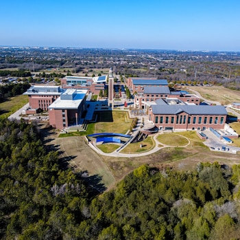 Vista aérea de un campus universitario con edificios de ladrillo, un reloj, césped, caminos y un área de asientos azules al aire libre, rodeado de árboles y vegetación