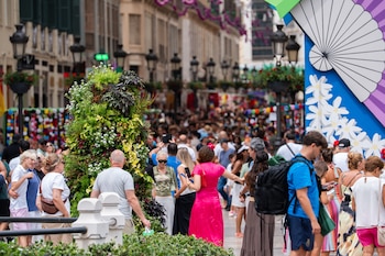 Una calle concurrida con edificios, farolas, plantas verdes, adornos coloridos, personas y una gran estructura decorativa blanca y azul