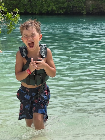 Un niño con chaleco salvavidas negro y bañador estampado camina en el agua clara cerca de una orilla con vegetación