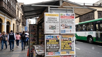 Un puesto de periódicos con periódicos exhibidos, personas caminando en la acera, edificios con balcones de madera, y un autobús verde y blanco en la calle